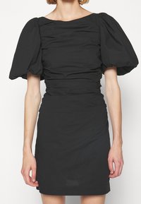 Svart klänning med fyrkantig halsringning, puffärmar och rynkad detalj vid midjan. Slätt tyg med en kroppsnära silhuett.