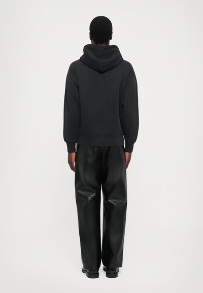 Un uomo si trova di spalle indossando una felpa con cappuccio nera, pantaloni di pelle neri larghi e scarpe nere su uno sfondo bianco uniforme.
