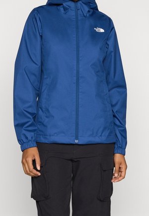 Blaues Kapuzenjacke mit Reißverschluss, elastischen Bündchen und The North Face Logo auf der linken Brust, getragen mit schwarzen Cargohosen.