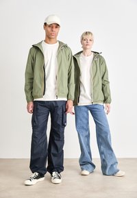 Due modelli indossano giacche leggere di colore verde oliva con cappucci, t-shirt beige, jeans cargo blu scuro e jeans svasati, completi di scarpe da ginnastica in tela.