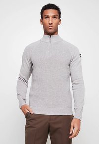 Hellgrauer Strickpullover mit hohem Kragen und halbem Reißverschluss. Verfügt über eine gerippte Struktur und kontrastierende braune Akzente an den Ärmeln.