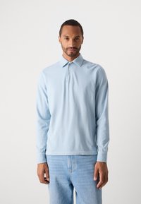 Mann med lyseblå langermet polo skjorte og lyseblå jeans, stående mot en enkel hvit bakgrunn.