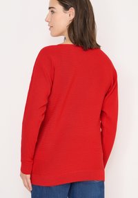Maglione rosso a coste con maniche lunghe e scollatura rotonda. Presenta un tessuto testurizzato e una vestibilità rilassata, che si estende leggermente oltre i fianchi.