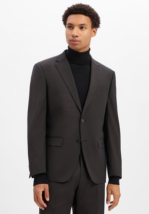 Mann trägt eine dunkle Anzugjacke und Hose mit einem schwarzen Rollkragenpullover, schaut leicht nach rechts auf weißem Hintergrund.
