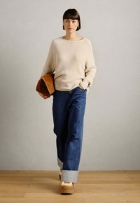 Beige ribbestrikket genser, avslappet passform, kombinert med høyt liv blå denimjeans med oppbrettede ender. Modell som holder en brun veske, tregulv.