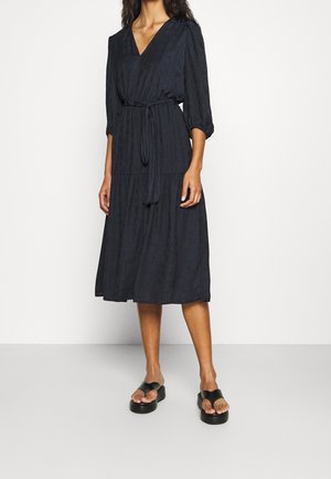 Kobieta w czarnej sukience midi z rękawami 3/4, dekoltem w serek i paskiem wiązanym w talii, do tego czarne sandały na platformie.