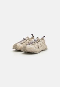 Beige joggesko med teksturert gummisåle, snøringsdesign, ventilasjonspaneler på siden og dragrem på hælen.