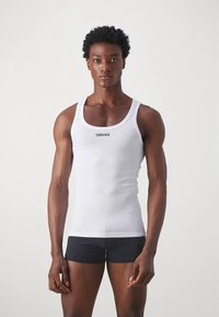 Ανδρικό μοντέλο που φοράει ένα εφαρμοστό λευκό φανελάκι Versace και μαύρα μποξερ σλιπ, στέκεται μπροστά από έναν απλό ανοιχτό φόντο.