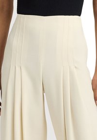 Korkeavyötäröiset kermaiset housut, joissa on pystysuuntaiset laskokset vyötäröllä, yhdistettynä sisätaskun mustaan ribbipaitaan.