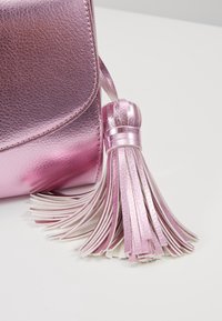Gros plan du coin d'un sac à main en cuir rose métallisé avec un grand pompon assorti sur une surface gris clair.