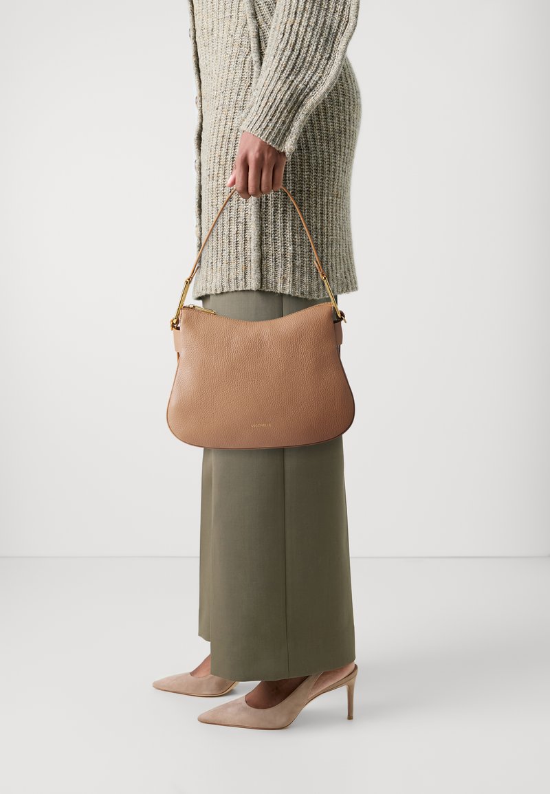 Borsa a tracolla in pelle color cuoio con forma curva, hardware dorato e manico. Abbinata a un maglione beige testurizzato e pantaloni verde oliva.