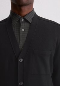 Schwarzer Cardigan mit V-Ausschnitt aus glattem Material, mit fünf Knöpfen und einer Brusttasche, getragen über einem dunklen karierten Hemd.