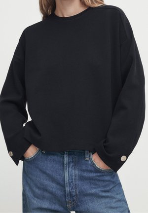 Femme portant un pull noir à manches longues avec des boutons argentés aux poignets et un jean en denim bleu taille haute, mains dans les poches.