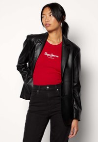 Giacca in pelle nera indossata sopra una maglietta rossa a maniche corte con logo bianco. Il modello abbina pantaloni neri a vita alta, che mettono in risalto una silhouette aderente.