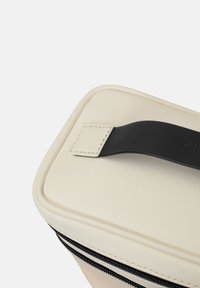 Trousse de maquillage rectangulaire en simili cuir crème avec une fermeture éclair noire et une poignée, présentant une texture lisse et des accents cousus.