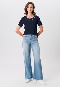 Marineblaues kurzärmliges T-Shirt mit dekorativen weißen Akzenten, kombiniert mit hellblauen weitgeschnittenen Jeans mit metallic floralem Stickmuster am Bund.