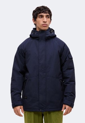 Mann trägt eine dunkelblaue Winterjacke mit Kapuze und Reißverschlusstaschen sowie grüne Hose, schaut seitlich vor einem schlichten Hintergrund.