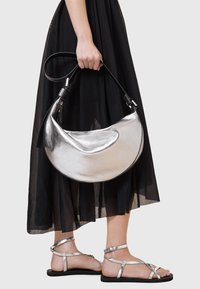 Borsa in pelle argentata metallizzata con forma curva a mezza luna e tracolla nera. Indossata con una gonna nera, trasparente e fluida, e sandali argentati.