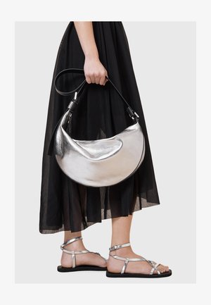 Metallisk silverfärgad läderväska med en böjd, halvmåneform och svart rem. Buren med en svart, genomskinlig, flödande kjol och silverfärgade sandaler.