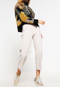 Strickpullover mit abstrakten gelben, grünen und grauen Mustern; kombiniert mit hellbeigen, maßgeschneiderten Hosen und metallischen Sneakern.