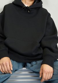 Sudadera negra con capucha hecha de tela gruesa con un bolsillo frontal, cierre con cordón y ajuste relajado. Combinada con pantalones azul claro.