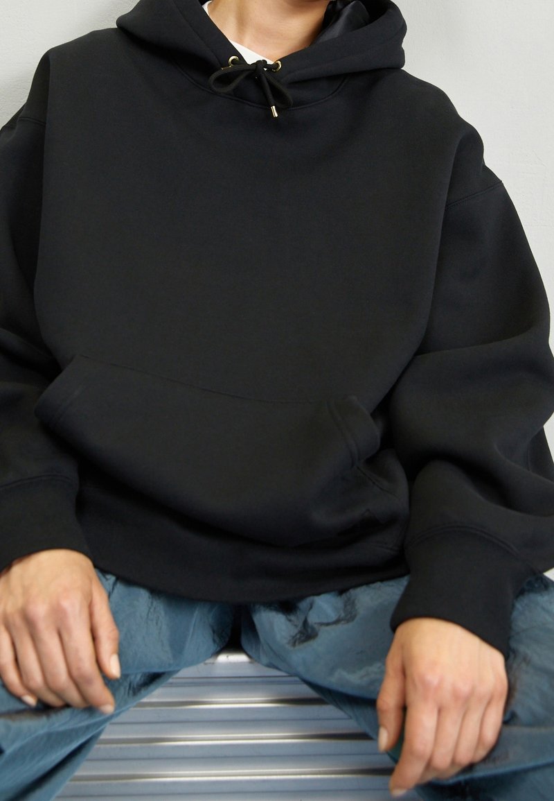 Sudadera negra con capucha hecha de tela gruesa con un bolsillo frontal, cierre con cordón y ajuste relajado. Combinada con pantalones azul claro.