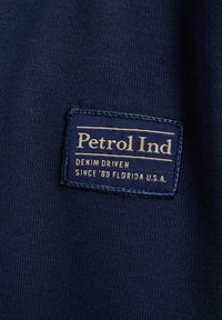 Material bleumarin cu o etichetă țesută care spune "Petrol Ind", având text auriu care afirmă "DENIM DRIVEN" și "SINCE '89 FLORIDA U.S.A."