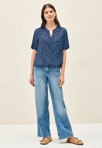 Blusa navy a maniche corte con motivo a foglie, scollo a V; abbinata a jeans a gamba larga azzurri e sandali beige con cinturino tra le dita.