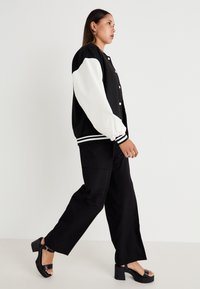 Veste de-varsité noire avec des manches blanches, rayures contrastantes, portée avec un pantalon large noir et des sandales à talons épais noires.