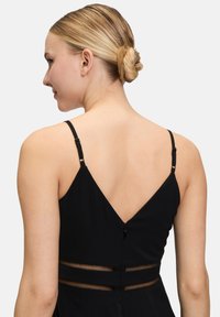 Zwarte jurk met dunne verstelbare banden, een diepe V-rug en gaasdetails bij de taille voor extra textuur en ontwerpcontrast.