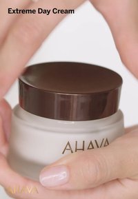 Vit krämbehållare med ett brunt lock, med guldfärgad text "AHAVA." Händer greppar behållaren, vilket framhäver dess runda form och släta yta.