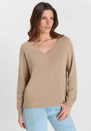 Femme aux cheveux bruns mi-longs portant un pull beige à col en V et un jean bleu clair, debout devant un fond blanc uni.