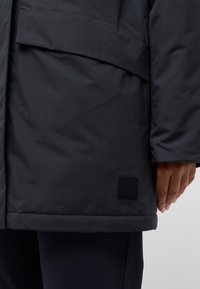 Donkergrijze geïsoleerde jas met een gladde textuur, grote voorzak en een zwart logo-opdruk aan de zoom. Getailleerde pasvorm bij de taille.