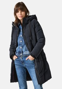 Abrigo acolchado negro con capucha, presenta un diseño acolchado y cierre frontal. Lleva encima una camisa de mezclilla y jeans. Manos en los bolsillos.