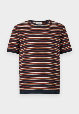 Homme debout les mains dans les poches portant une chemise à manches courtes rayée, un short beige avec une ceinture noire et des mocassins noirs sur fond blanc.