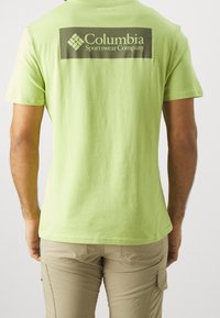 Světle zelené tričko s krátkým rukávem vyrobené z bavlny, s tmavohnědým logem společnosti Columbia Sportswear na zadní straně.