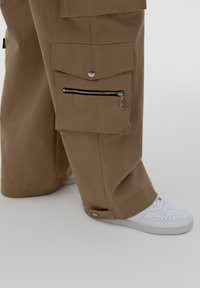 Zakkige, bruine cargobroeken hebben meerdere zakken, een ritszak aan de zijkant en een drukknoopsluiting aan de broekspijp, gecombineerd met witte sneakers.