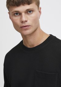 Black waffle-knit long-sleeve shirt with rounded neckline and a chest pocket. Smooth texture and subtle stitching details.