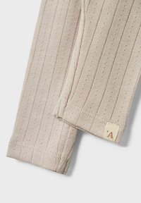 Vêtement en tricot beige avec une texture côtelée, présentant des lignes verticales et de petits trous. Étiquette avec la lettre 'A' visible sur une couture.