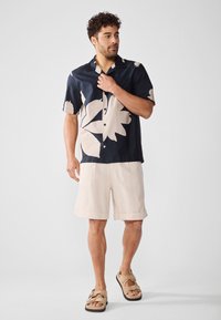Camisa de manga corta navy con grandes patrones de hojas beige, combinada con pantalones cortos de lino beige. El modelo lleva sandalias marrones con doble tira.