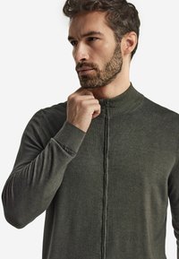 Ciemnozielona bluza na zamek z wysokim kołnierzem, długimi rękawami i ribbowymi mankietami. Gładka tekstura materiału z minimalnymi detalami stębnowania.