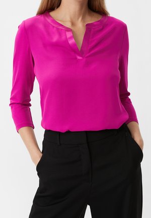 Femme portant un chemisier rose vif à manches longues avec un col en V, associé à un pantalon noir taille haute, mains dans les poches.