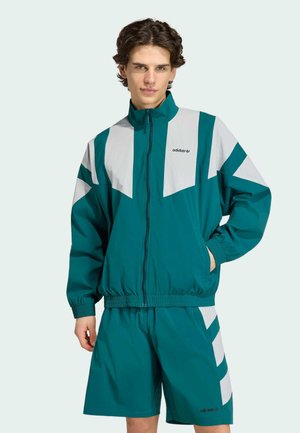 Ung mand iført teal og lysegrå Adidas træningsjakke og matchende shorts, hænderne i lommerne, stående mod en ensfarvet lys baggrund.