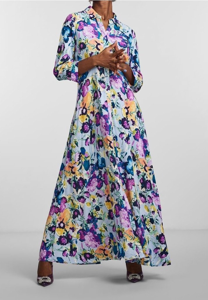 Femme portant une longue robe à boutons à fleurs avec des fleurs violettes, jaunes et bleues, assortie à des talons violets ornés de bijoux.
