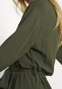 Olivgrön långärmad topp med en knytning i midjan, tillverkad av ett mjukt, lätt tyg, med mjuka draperingar och en slät yta.