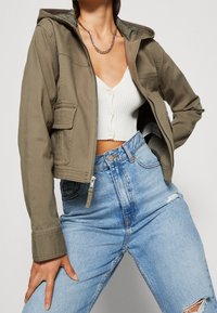 Person som bär en olivgrön zip-up jacka, vit räfflad knäppbar crop top, silverkedja och hög midja ljusblå ripped jeans.