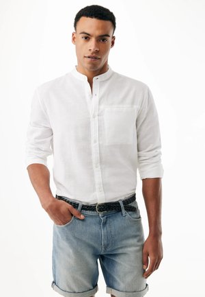 Mężczyzna ubrany w białą koszulę na guziki z podwiniętymi rękawami, jasnoniebieskie szorty z dżinsu oraz czarny pleciony pasek, stojący na białym tle.