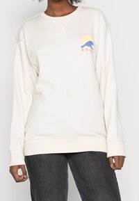Sudadera de color crema con puños y dobladillo acanalados, con un logotipo gráfico en tonos pastel que muestra un diseño de sol y ola en el área del pecho izquierdo.