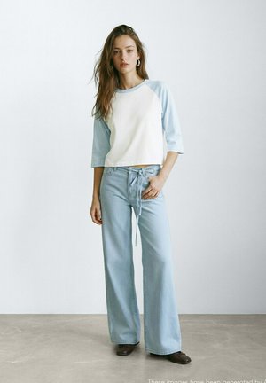 Mlada ženska stoji v notranjosti, oblečena v belo in svetlomodro dolgoročno majico ter svetlomodre široke kavbojke z rjavimi čevlji.