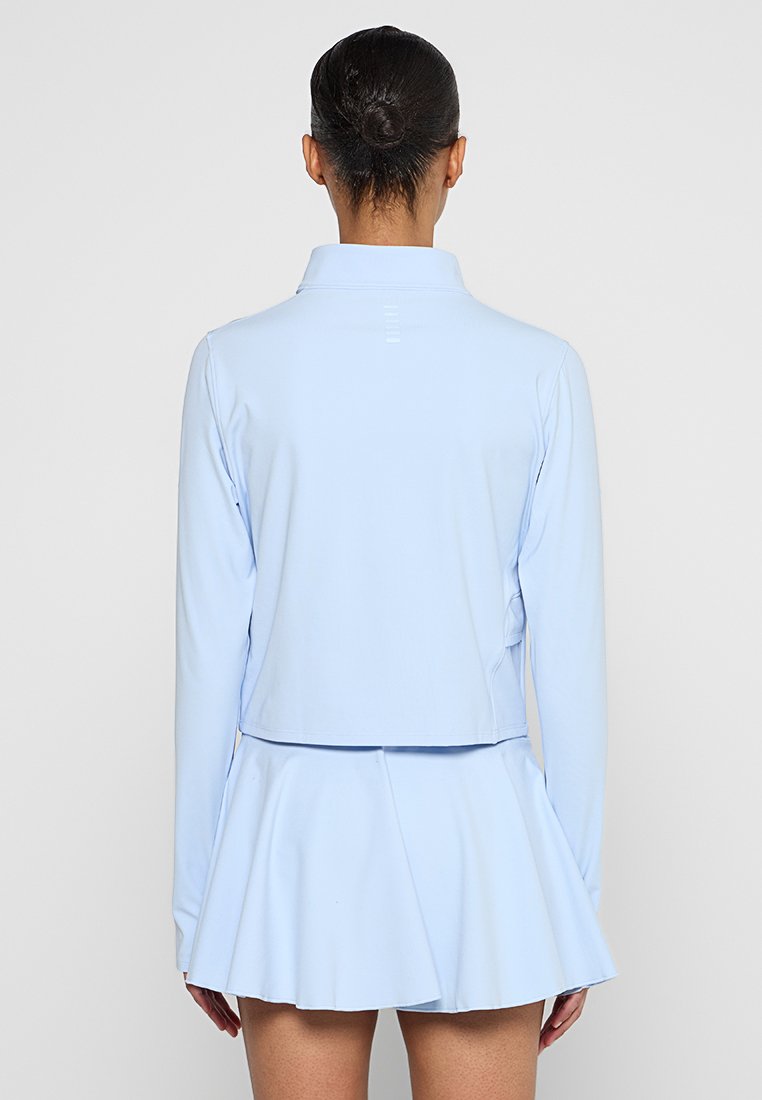 Femme avec un chignon foncé portant un haut à manches longues bleu clair et une jupe plissée, se tenant dos à la caméra devant un fond uni.